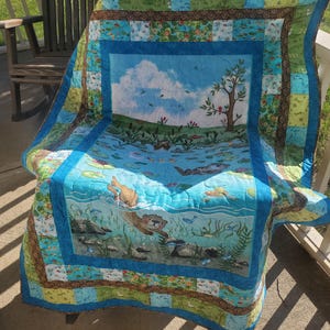 May include: A colorful quilt featuring a central panel with an otter swimming in a pond, surrounded by fish and aquatic plants. The quilt has a blue border and patchwork design with green, blue, and brown squares. The quilt is draped over a chair.