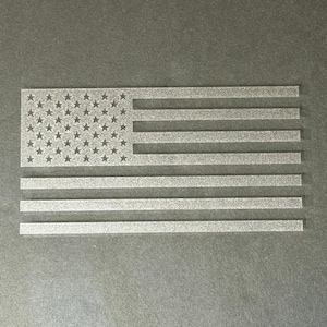 May include: A silver-colored American flag design with stars and stripes. The flag is cut out of a solid material and is displayed against a gray background. The stars are in the upper left corner.