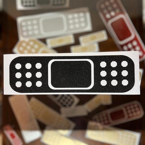 May include: Black and white bandage sticker with a glittery texture. The sticker has a rectangular center and a series of white dots on each side. Other bandage stickers in various colors are in the background.