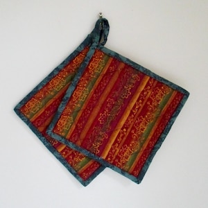May include: Two square quilted pot holders with a teal blue border and hanging loop. The pot holders feature a striped pattern in shades of red, orange, and green with a floral design.
