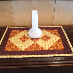 May include: A white hobnail vase sits on a handmade quilted table runner. The runner features a geometric design with shades of orange, yellow, and brown. The runner is rectangular and has a brown border.