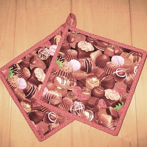 May include: Two square pot holders with a repeating pattern of various chocolates in shades of brown, pink, and white. The pot holders have a red border and a loop for hanging. The background is a light wood surface.