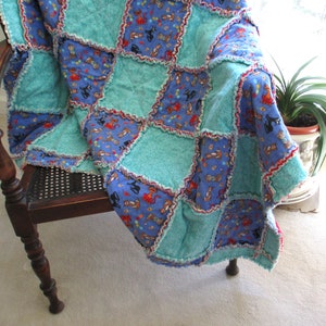 May include: A rag quilt featuring a checkerboard pattern of solid teal fabric squares and blue fabric squares printed with a repeating pattern of colorful cats. The quilt has exposed frayed seams between each square and is draped over a wooden chair.
