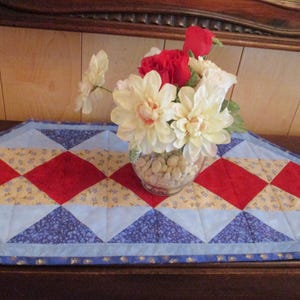 May include: A quilted table runner with a patriotic design. The runner features red, white, and blue diamond and triangle patterns. A vase of white, red, and cream-colored flowers sits on the runner.