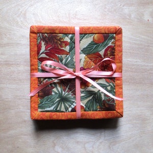 May include: A set of four orange fabric coasters with a floral pattern and a pink ribbon tied around them.