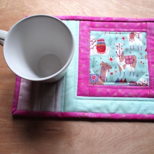 May include: A pink and blue quilted coaster with a pattern of llamas and cacti. The coaster is sitting on a wooden surface with a white mug on top of it.