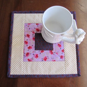 May include: A white ceramic mug with a decorative handle rests on a square quilted coaster. The coaster features a patchwork design with floral and polka dot patterns in shades of purple, pink, and cream.