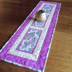 May include: A quilted table runner with a floral pattern in shades of purple, green, and cream. The runner features a central panel with floral designs, framed by cream and purple borders. A gold-colored metal candle holder sits on top.