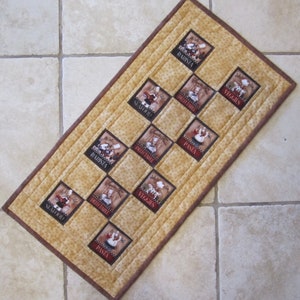 May include: A rectangular quilted table runner with a tan speckled fabric background and a dark brown border. It features ten square patches depicting chefs with the text Barista, Seafood, Delectable, Veggies, and Pasta printed on each square.