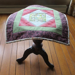 May include: A square quilted fabric topper featuring a central panel with a multi-colored animal print, surrounded by white, red, and green geometric borders, and finished with a dark purple outer border patterned with small black leaf silhouettes.