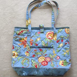 May include: A quilted tote bag with a floral pattern in shades of blue, red, yellow, and green. The bag has two handles, a small pocket, and a button closure. The bag is approximately 14 inches wide and 12 inches tall.