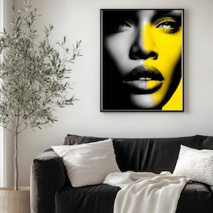 Może przedstawiać: Oprawiony wydruk artystyczny przedstawiający portret kobiety, podzielony pionowo na czarno-biały i żółty. Usta kobiety są ciemne, a jej oczy są podkreślone żółtym kolorem. Grafika jest prezentowana nad czarną sofą.