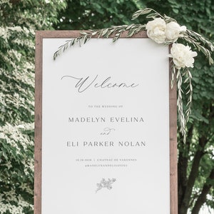 May include: A wooden welcome sign with a white background and black text. The text reads "Welcome to the wedding of Madelyn Evelina and Eli Parker Nolan 10.26.2028 Chateau de Varennes #MadelynAndEliSayOui".