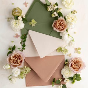 May include: Three envelopes in different shades of green, beige, and brown are arranged in a stack. The envelopes are surrounded by white and pink flowers and greenery.