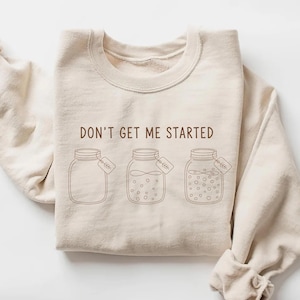 May include: A beige sweatshirt with the phrase "DON'T GET ME STARTED" printed in brown. Below the text are three brown line-drawn mason jars, each with a small tag. The jars contain different fillings.