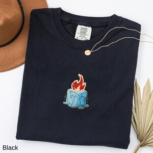 May include: Black crew neck t-shirt with a candle graphic. The candle is blue with red flames and dripping wax. A gold necklace and a brown hat are also visible.