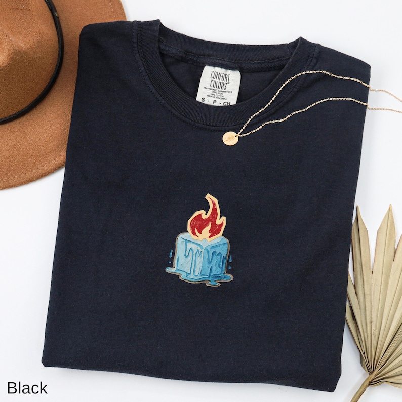 May include: Black crew neck t-shirt with a candle graphic. The candle is blue with red flames and dripping wax. A gold necklace and a brown hat are also visible.