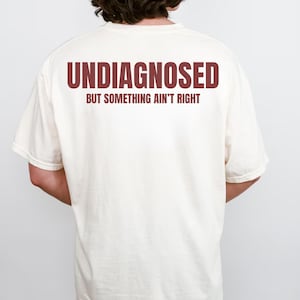 May include: Cream-colored t-shirt with the text "UNDIAGNOSED" in maroon block letters above "BUT SOMETHING AIN'T RIGHT". The shirt is a casual style, with short sleeves and a relaxed fit. The text is centered on the back of the shirt.