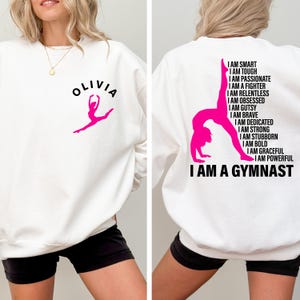 Puede incluir: Sudadera blanca con un gráfico de gimnasta rosa y el nombre "OLIVIA" en la parte delantera. La parte trasera presenta un gráfico de gimnasta rosa y el texto "I AM A GYMNAST" junto con afirmaciones empoderadoras como "I AM STRONG" y "I AM BRAVE".