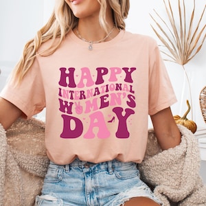 Op de afbeelding: Een roze T-shirt met de tekst "Happy International Women's Day" in roze en paarse letters met een vredesteken en een afbeelding van een hoge hak.