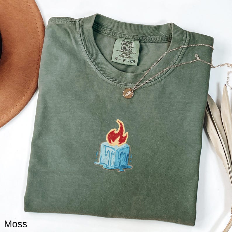 May include: A moss green Comfort Colors t-shirt with a graphic of a blue ice cube with red flames. A gold necklace with a pendant is on the shirt. The shirt is labeled "S - P - CH".