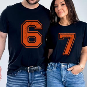 May include: Two black t-shirts with orange numbers 6 and 7. The numbers have a distressed, web-like design inside and a black outline. The shirts are paired with blue jeans. The image shows a couple wearing the shirts.
