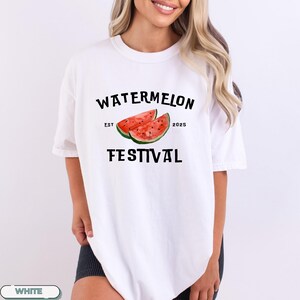 Pu&ograve; includere: T-shirt bianco con un design a tema anguria. La grafica include le parole "WATERMELON FESTIVAL" con l'anno "2025" e due fette di anguria. La maglietta &egrave; uno stile casual a maniche corte.