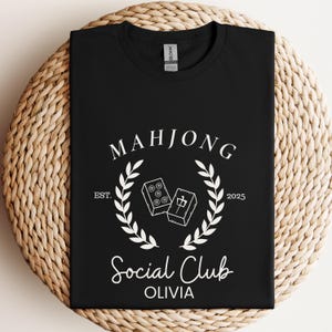May include: Black t-shirt with white text that reads "Mahjong Social Club Olivia" with a wreath design and the text "Est. 2025".