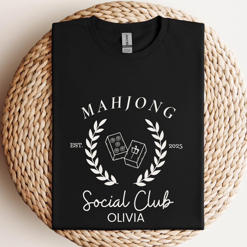 May include: Black t-shirt with white text that reads "Mahjong Social Club Olivia" with a wreath design and the text "Est. 2025".