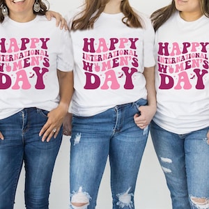 Op de afbeelding: Drie witte T-shirts met de tekst "Happy International Women's Day" in roze en bordeauxrode letters. De shirts worden gedragen door drie personen.