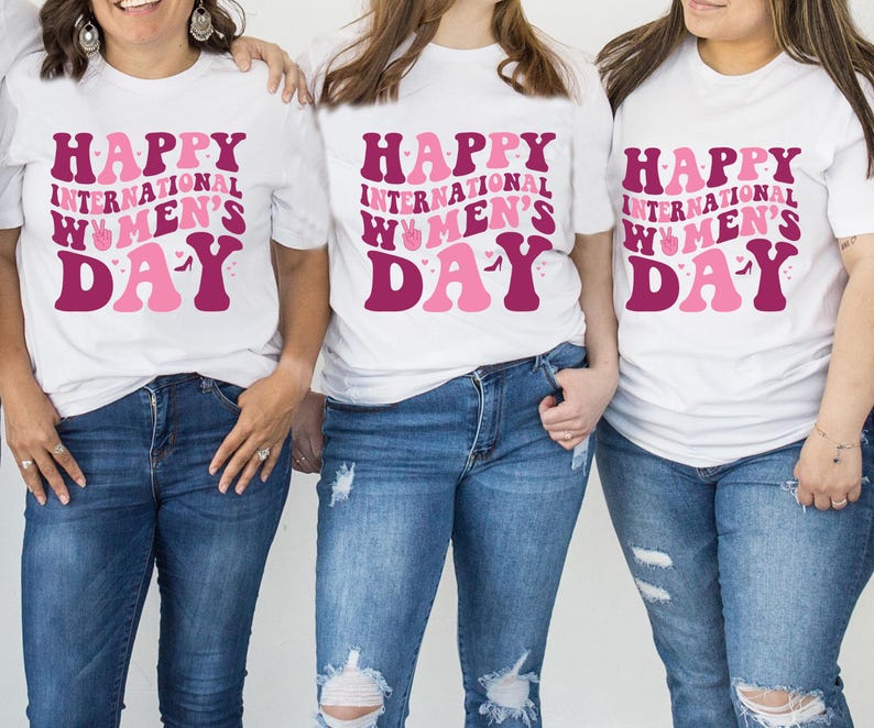 Op de afbeelding: Drie witte T-shirts met de tekst "Happy International Women's Day" in roze en bordeauxrode letters. De shirts worden gedragen door drie personen.