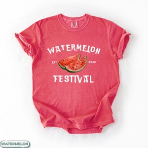Pu&ograve; includere: Una maglietta rossa con le parole "WATERMELON FESTIVAL" in bianco. La maglietta presenta un'illustrazione ad acquerello di due fette di anguria. Il testo "EST 2025" &egrave; anch'esso stampato.