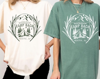 May include: Two women wearing white and green t-shirts with a graphic design of a campsite and the text "AUGUST 2025 Garcia's Bachelorette CAMP BACH SEDONA, AZ"