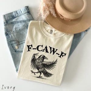 May include: Ivory-colored t-shirt with a black graphic of a crow and the text "F-CAW-F". The shirt is paired with light blue jeans, a tan hat, and a necklace. The shirt is made by Comfort Colors.