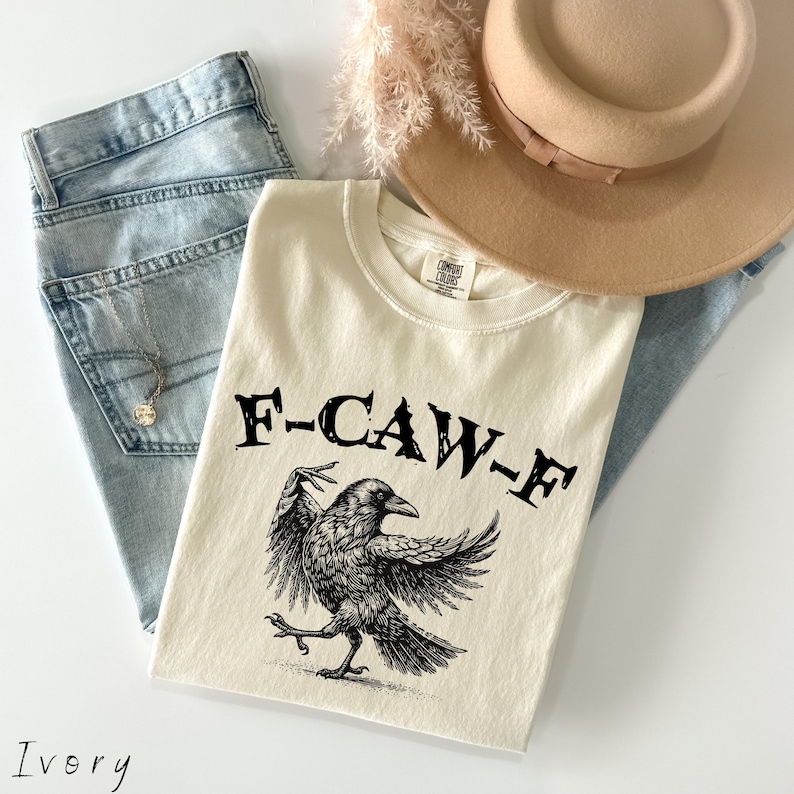 May include: Ivory-colored t-shirt with a black graphic of a crow and the text "F-CAW-F". The shirt is paired with light blue jeans, a tan hat, and a necklace. The shirt is made by Comfort Colors.