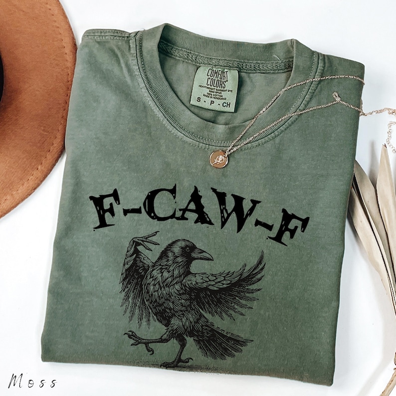 May include: Moss green t-shirt with the text "F-CAW-F" above a black crow illustration. The shirt has a crew neck and short sleeves. A gold necklace with a pendant is draped across the shirt.