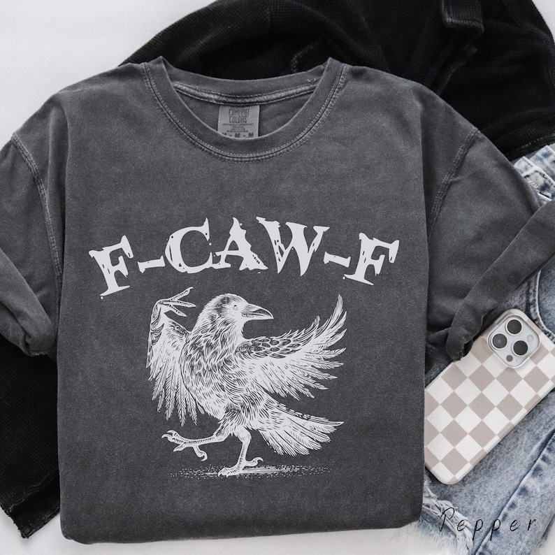 May include: Dark gray sweatshirt with the text "F-CAW-F" above a detailed white illustration of a crow with outstretched wings. The sweatshirt has a crew neck and long sleeves. A phone case with a checkered pattern is visible.