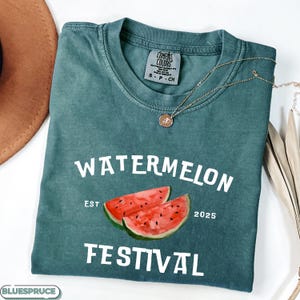 Pu&ograve; includere: Una maglietta Comfort Colors verde acqua con le parole "WATERMELON FESTIVAL" in bianco. La maglietta presenta un'illustrazione ad acquerello di due fette di anguria. Una collana d'oro con un ciondolo &egrave; drappeggiata sulla maglietta.