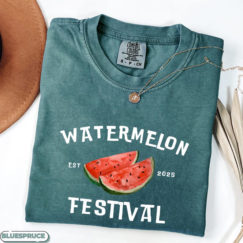 Pu&ograve; includere: Una maglietta Comfort Colors verde acqua con le parole "WATERMELON FESTIVAL" in bianco. La maglietta presenta un'illustrazione ad acquerello di due fette di anguria. Una collana d'oro con un ciondolo &egrave; drappeggiata sulla maglietta.