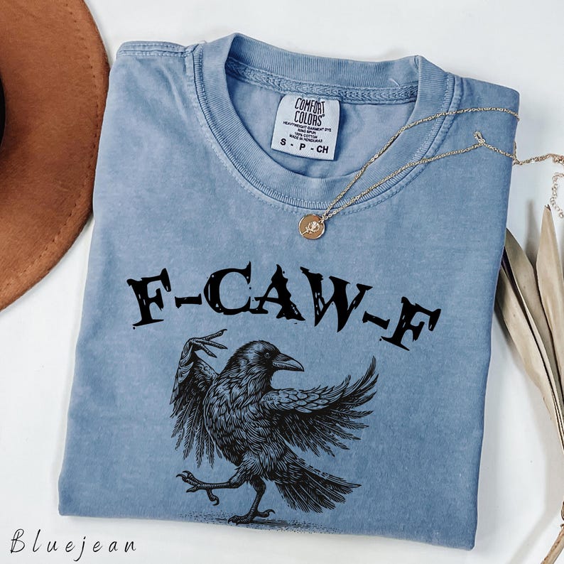 May include: A light blue Comfort Colors t-shirt with the text "F-CAW-F" above a detailed black raven illustration. A gold necklace with a pendant is draped across the shirt. The shirt is labeled S-P-CH.