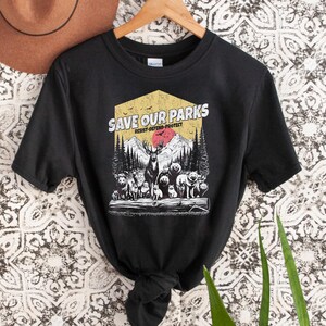May include: Black t-shirt with a vintage-style graphic that says "Save Our Parks" with a red sun, mountains, and animals. The text below the graphic says "Resist-Defend-Protect".
