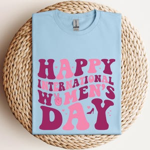 Op de afbeelding: Lichtblauw T-shirt met de tekst "Happy International Women's Day" in roze en bordeauxrode letters. De tekst is in een retro-lettertype stijl. Het shirt heeft een vredesteken met een hart in het midden.