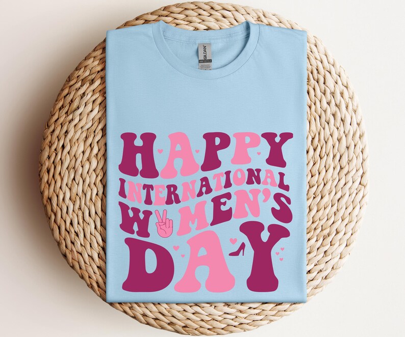 Op de afbeelding: Lichtblauw T-shirt met de tekst "Happy International Women's Day" in roze en bordeauxrode letters. De tekst is in een retro-lettertype stijl. Het shirt heeft een vredesteken met een hart in het midden.