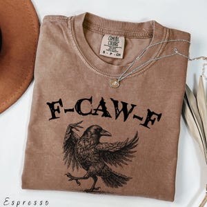 May include: Espresso-colored t-shirt with the text "F-CAW-F" above a detailed illustration of a crow. The shirt has a crew neck and short sleeves. A silver necklace with a pendant is draped across the shirt.