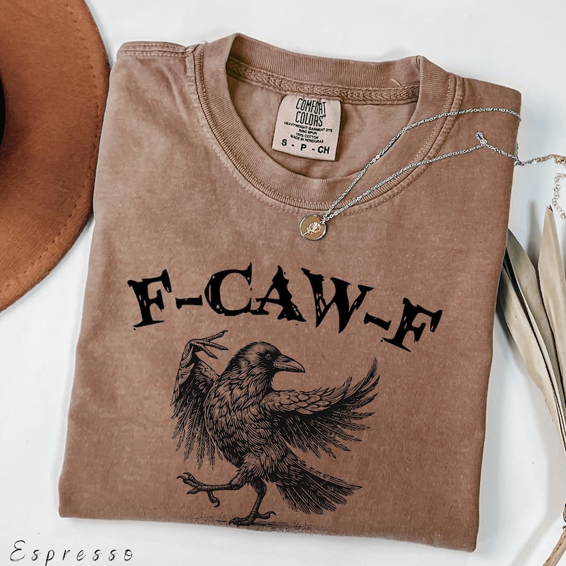 May include: Espresso-colored t-shirt with the text "F-CAW-F" above a detailed illustration of a crow. The shirt has a crew neck and short sleeves. A silver necklace with a pendant is draped across the shirt.