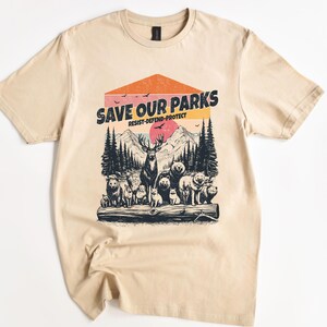 May include: A beige t-shirt with a vintage-style graphic print of a forest scene with animals. The text "Save Our Parks" is printed in black with the words "Resist-Defend-Protect" underneath.