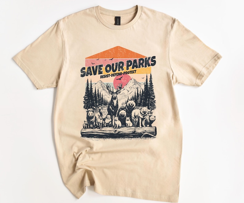 May include: A beige t-shirt with a vintage-style graphic print of a forest scene with animals. The text "Save Our Parks" is printed in black with the words "Resist-Defend-Protect" underneath.