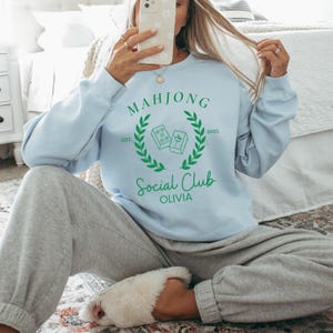 May include: A light blue sweatshirt with a green graphic design that says "Mahjong Social Club Olivia" with a wreath and the text "EST 2025".
