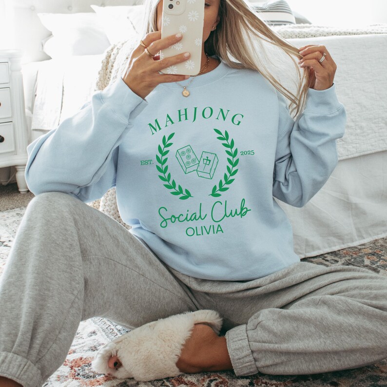 May include: A light blue sweatshirt with a green graphic design that says "Mahjong Social Club Olivia" with a wreath and the text "EST 2025".