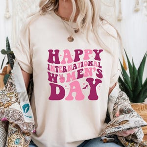 Op de afbeelding: Een wit T-shirt met de tekst "Happy International Women's Day" in roze en paarse letters. De letters zijn gestileerd met harten en een vredesteken.